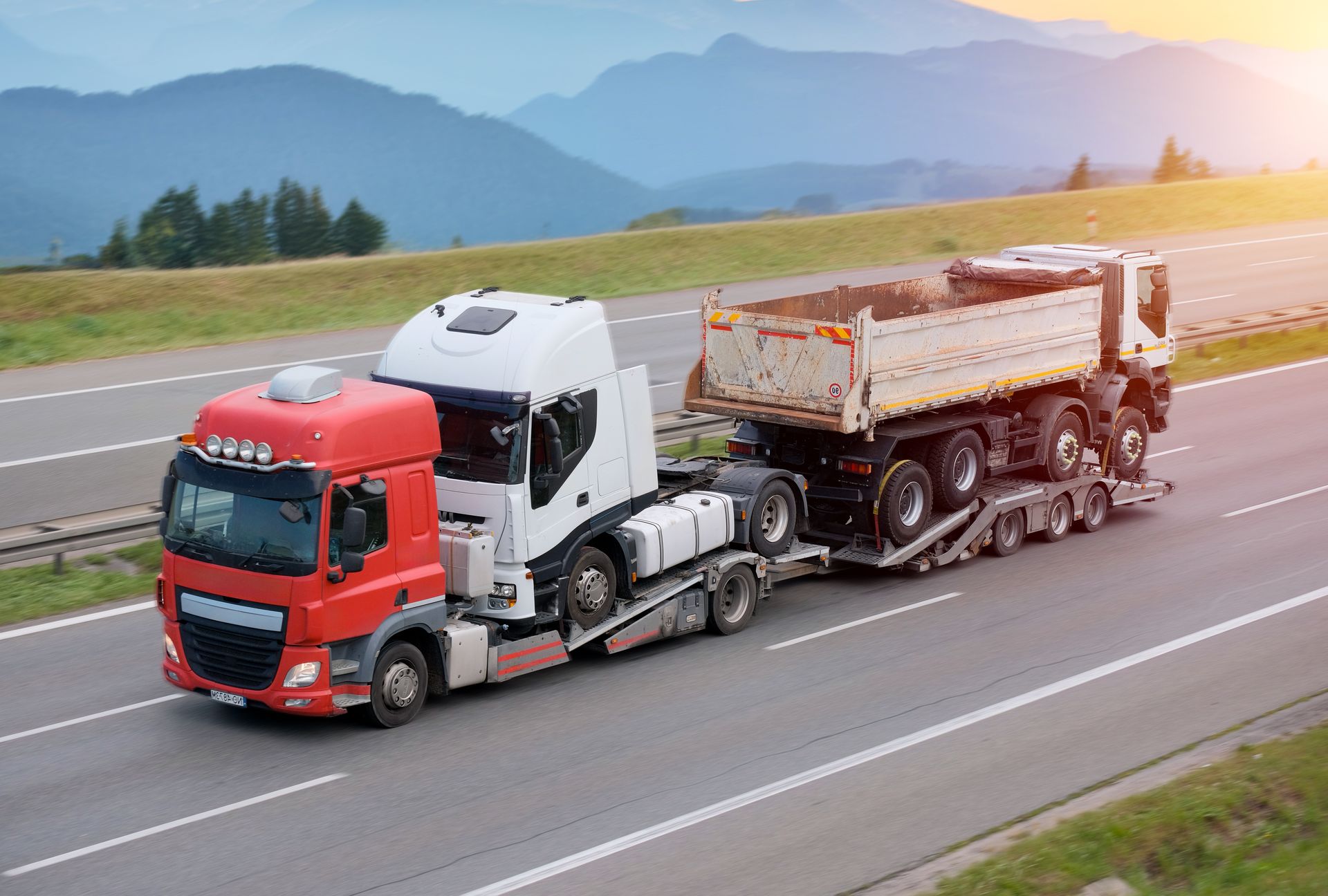 Truck hauling two heavy vehicles on a highway through a hilly landscape.