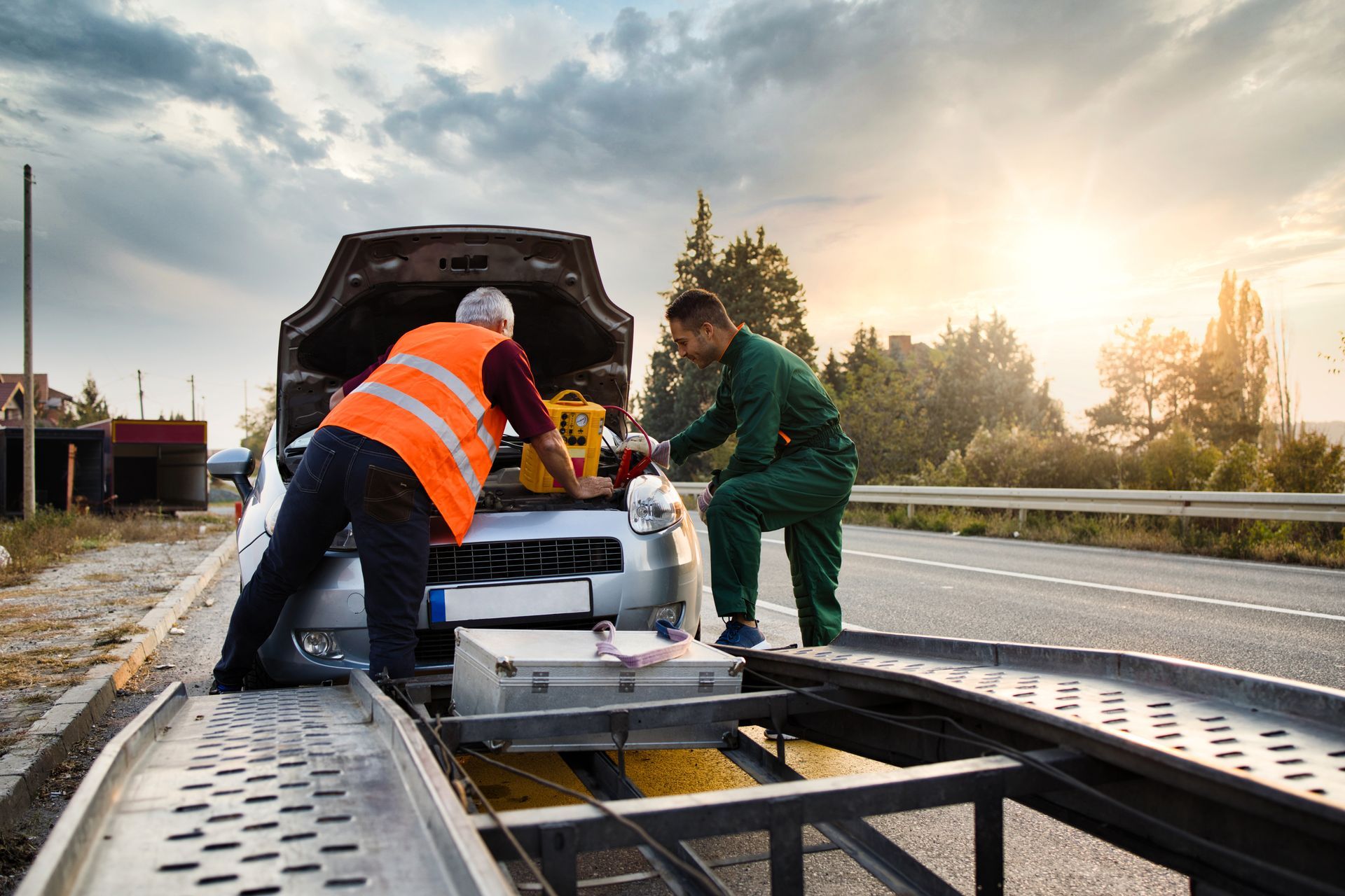 Two workers assist a broken-down car on the roadside before loading it onto a tow truck.