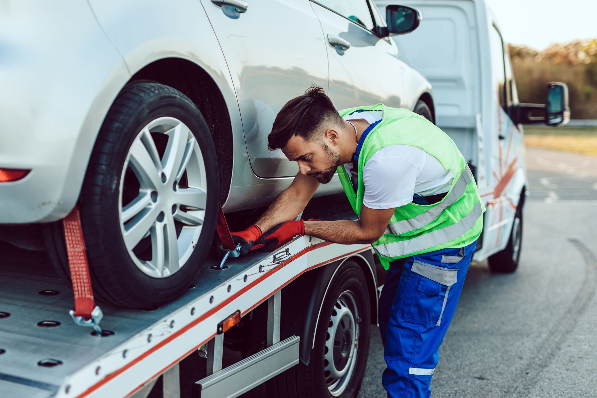 Tow truck operator securing a car to the flatbed. The man wears a safety vest, blue pants, and is focused on the task. Tow truck operator securing a car to the flatbed. The man wears a safety vest, blue pants, and is focused on the task.