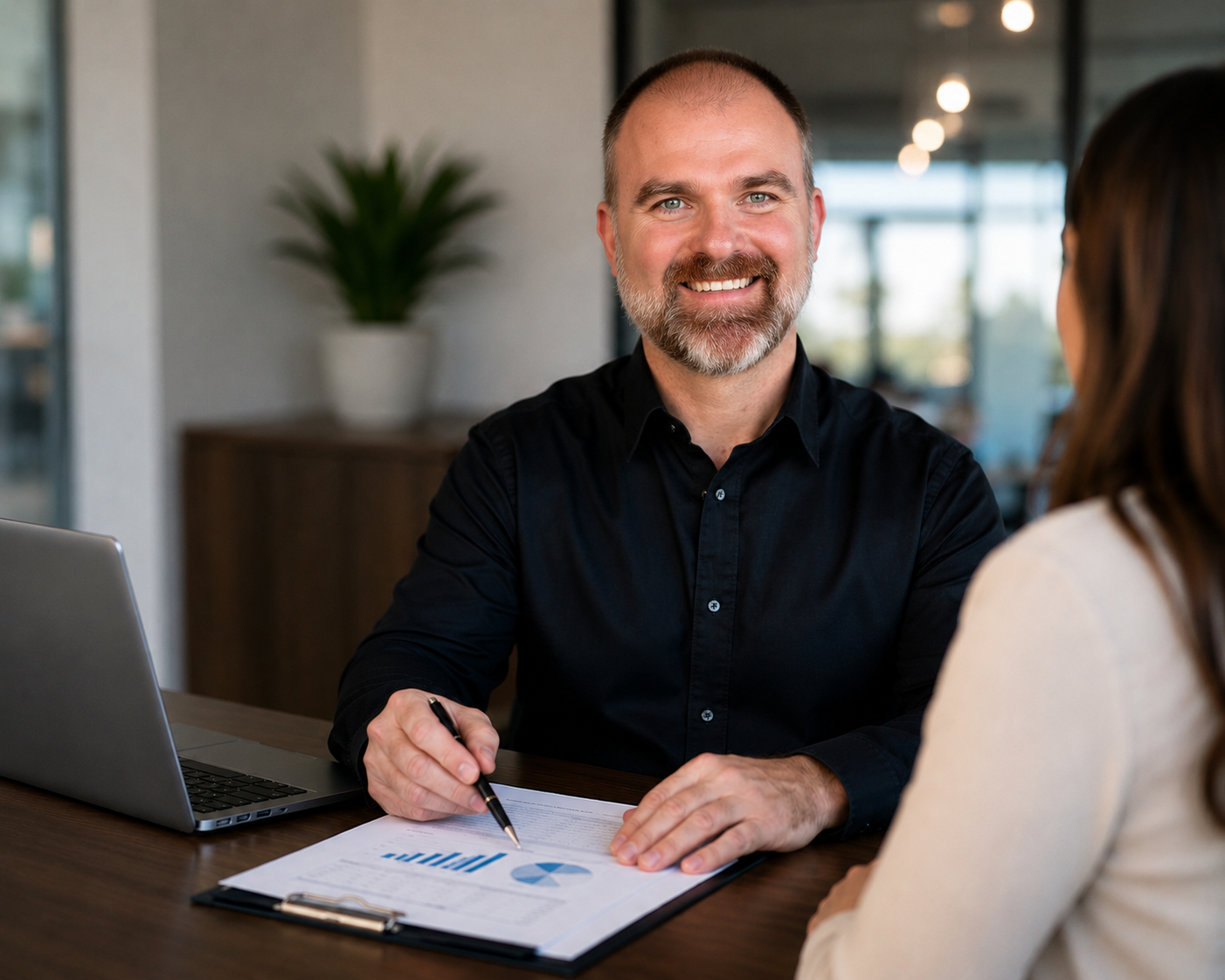 Smiling man in a black shirt reviewing charts with a woman across a desk in an office