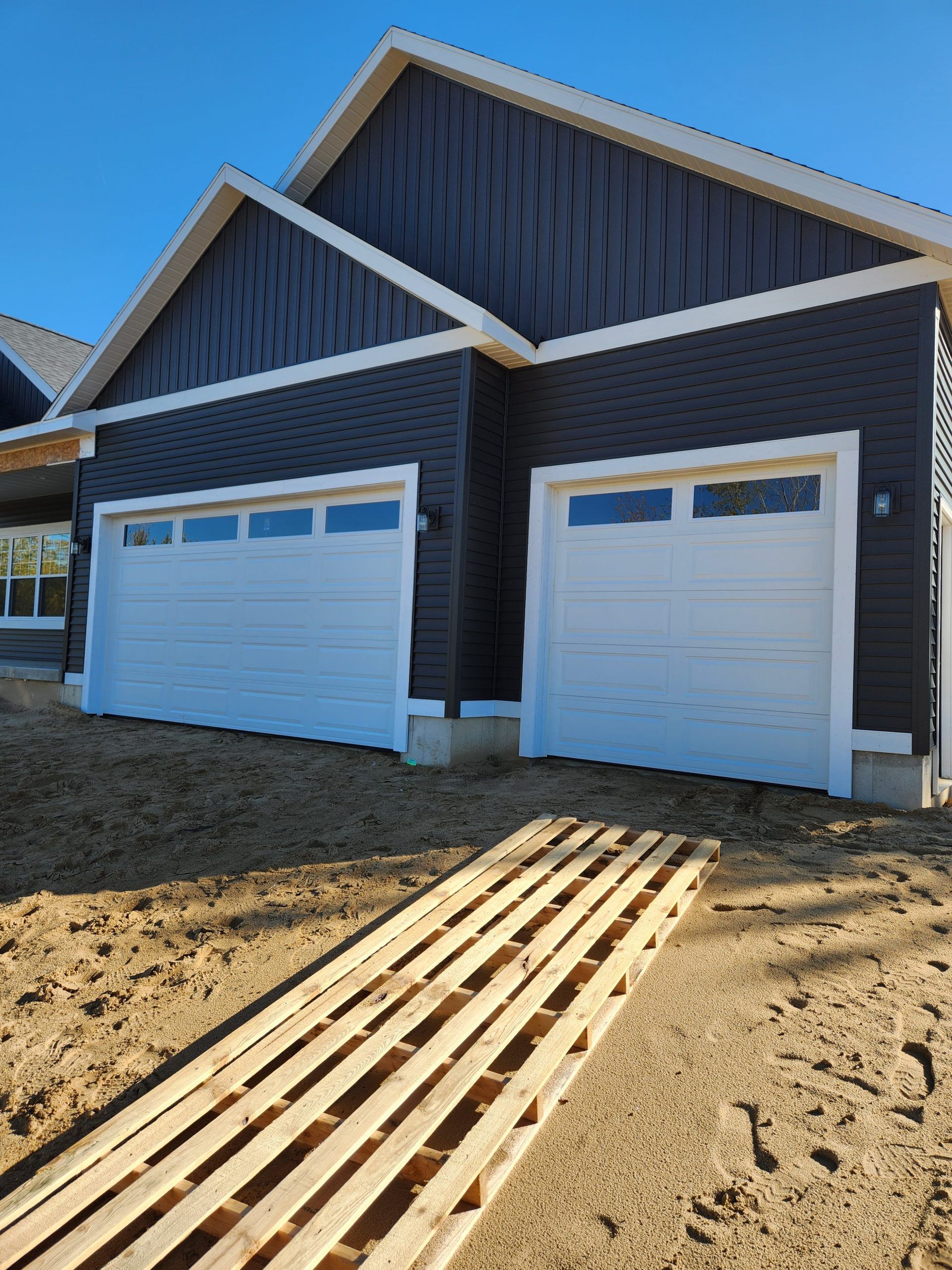 Two-car garage with white doors, dark gray siding, and a wooden ramp on a dirt lot under a blue sky.