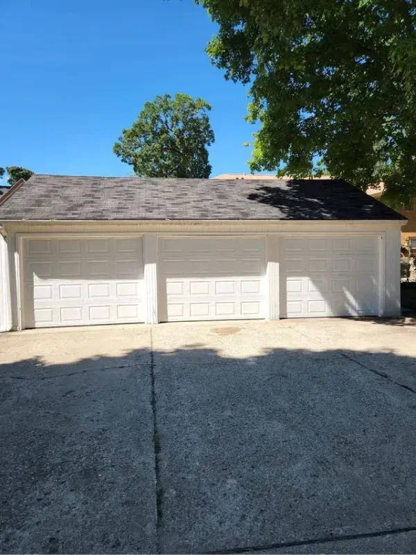 Three-car garage with white doors under a dark roof, on a sunny day. Concrete driveway in front.