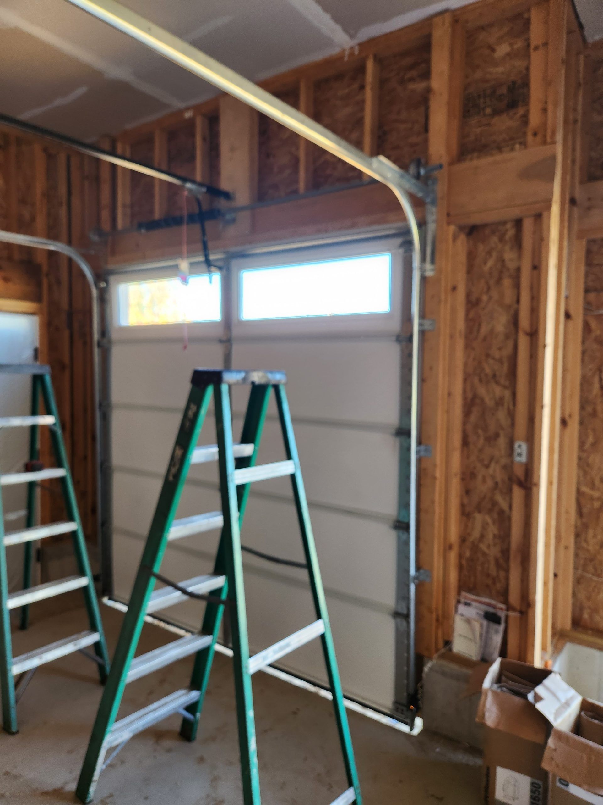 Interior view of a garage with a partially installed garage door. Two ladders are in front of the closed door.