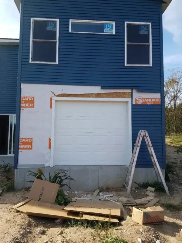 Blue house under construction; white garage door, windows, ladder. Cardboard and debris on ground.