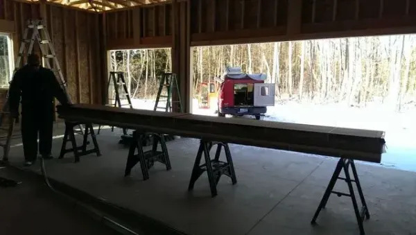 Man inside a wood-framed structure, working on long beam supported by sawhorses. Winter scene visible through windows.