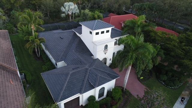 An aerial view of a large white house with a black roof surrounded by palm trees.