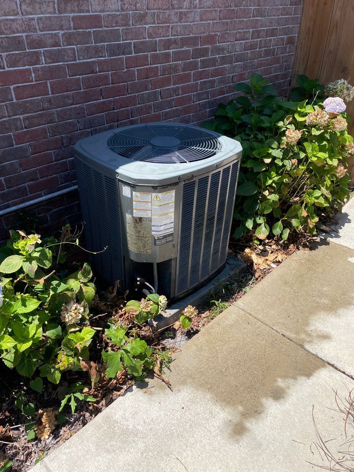 An outdoor gray HVAC condenser unit positioned next to a brick wall and a flowering hydrangea bush.