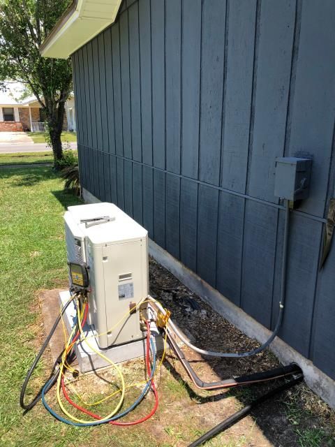An outdoor HVAC unit next to a dark gray house wall with connected technician hoses and electrical wiring.