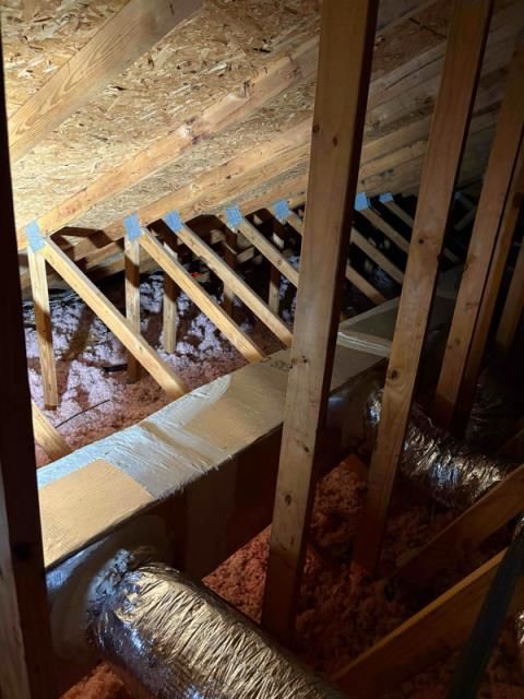 A view inside an attic showing wooden roof trusses, foil-wrapped air ducts, and pink insulation between ceiling joists.