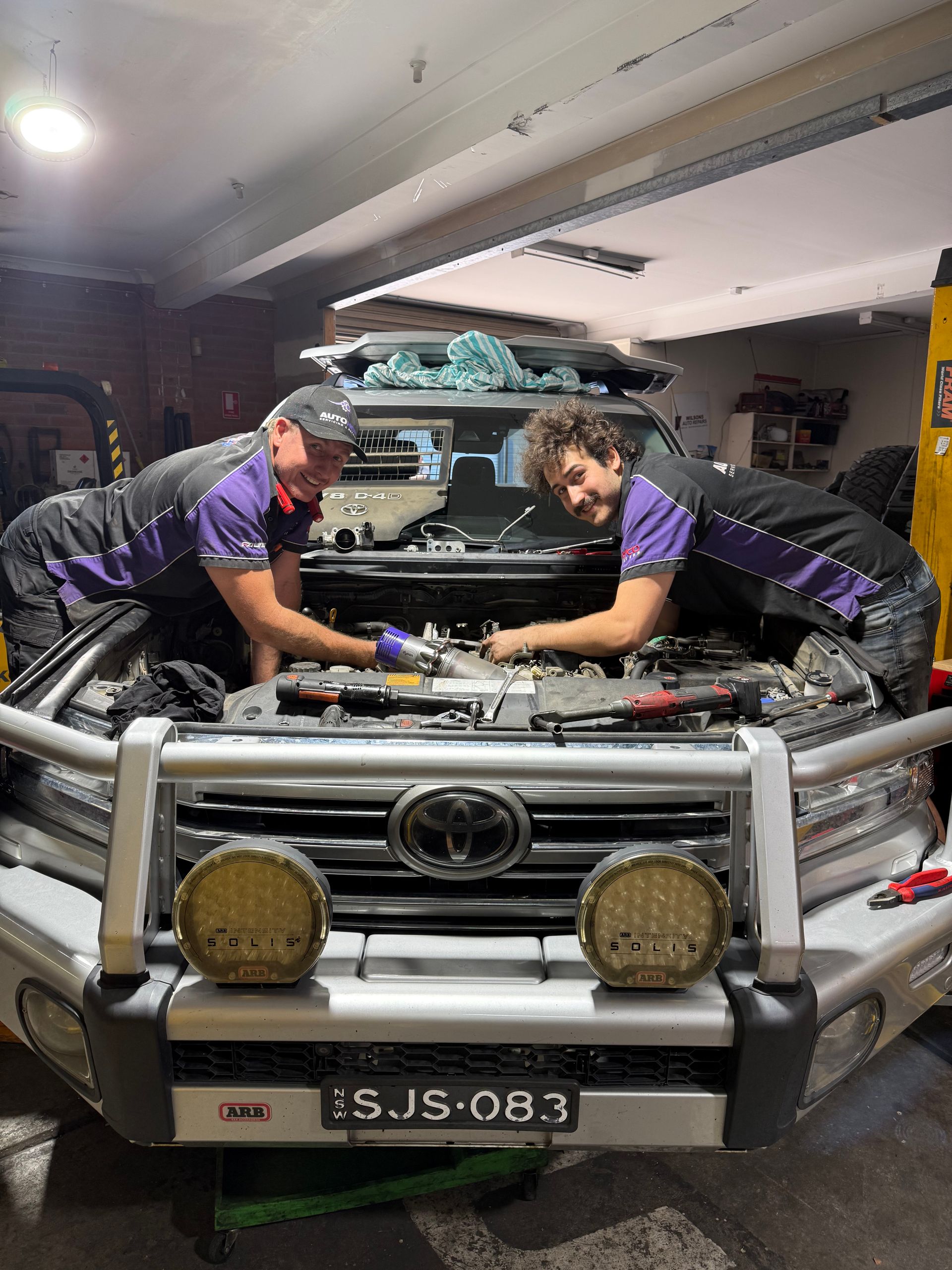 Two mechanics in a garage working on a vehicle's engine, wearing purple shirts, with tools visible.— AutoHop Lambton in Lambton, NSW