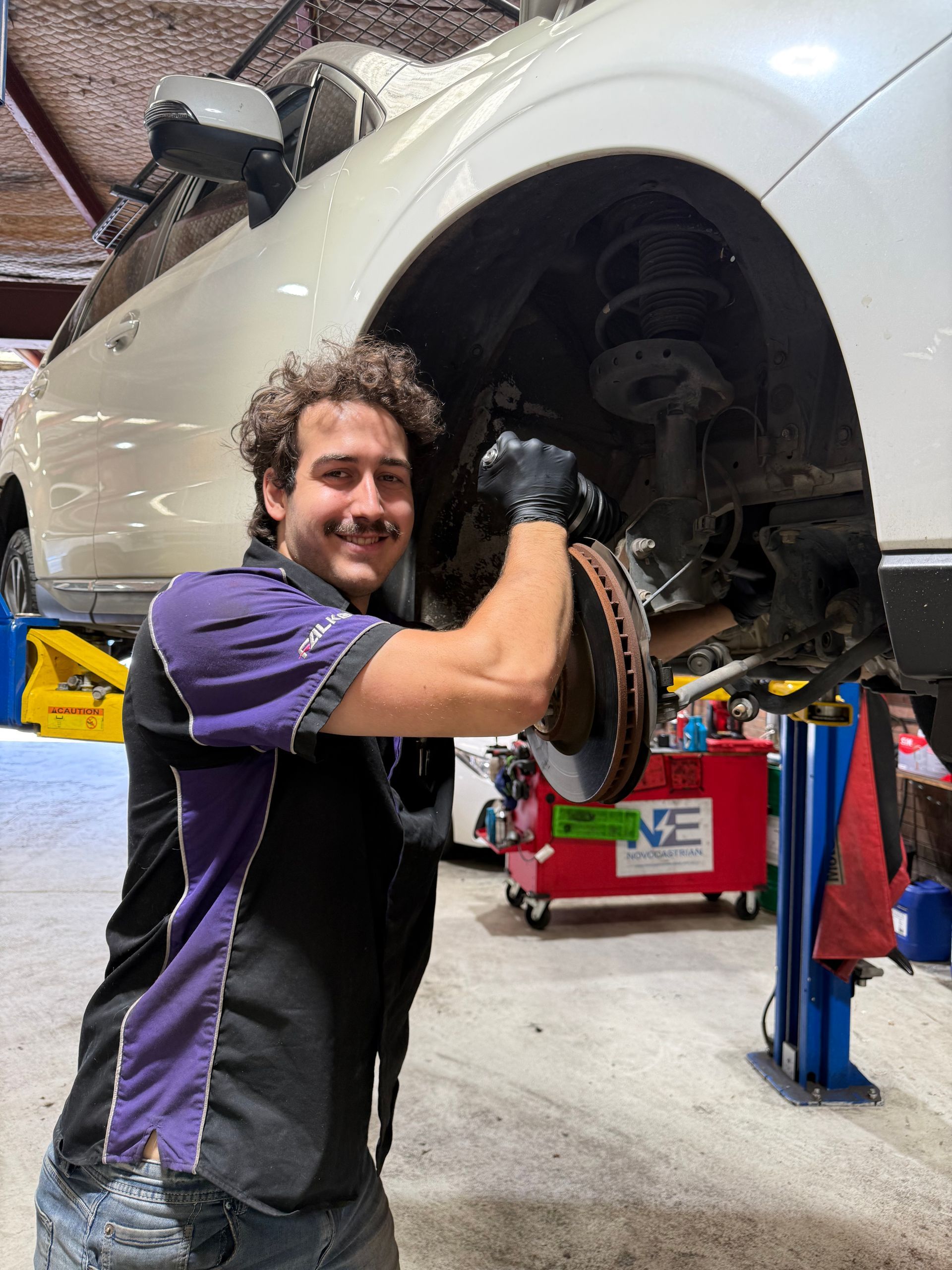 Mechanic holding a car brake rotor in a garage. Wearing gloves, black and purple shirt, smiling. Car on a lift.— AutoHop Lambton in Lambton, NSW