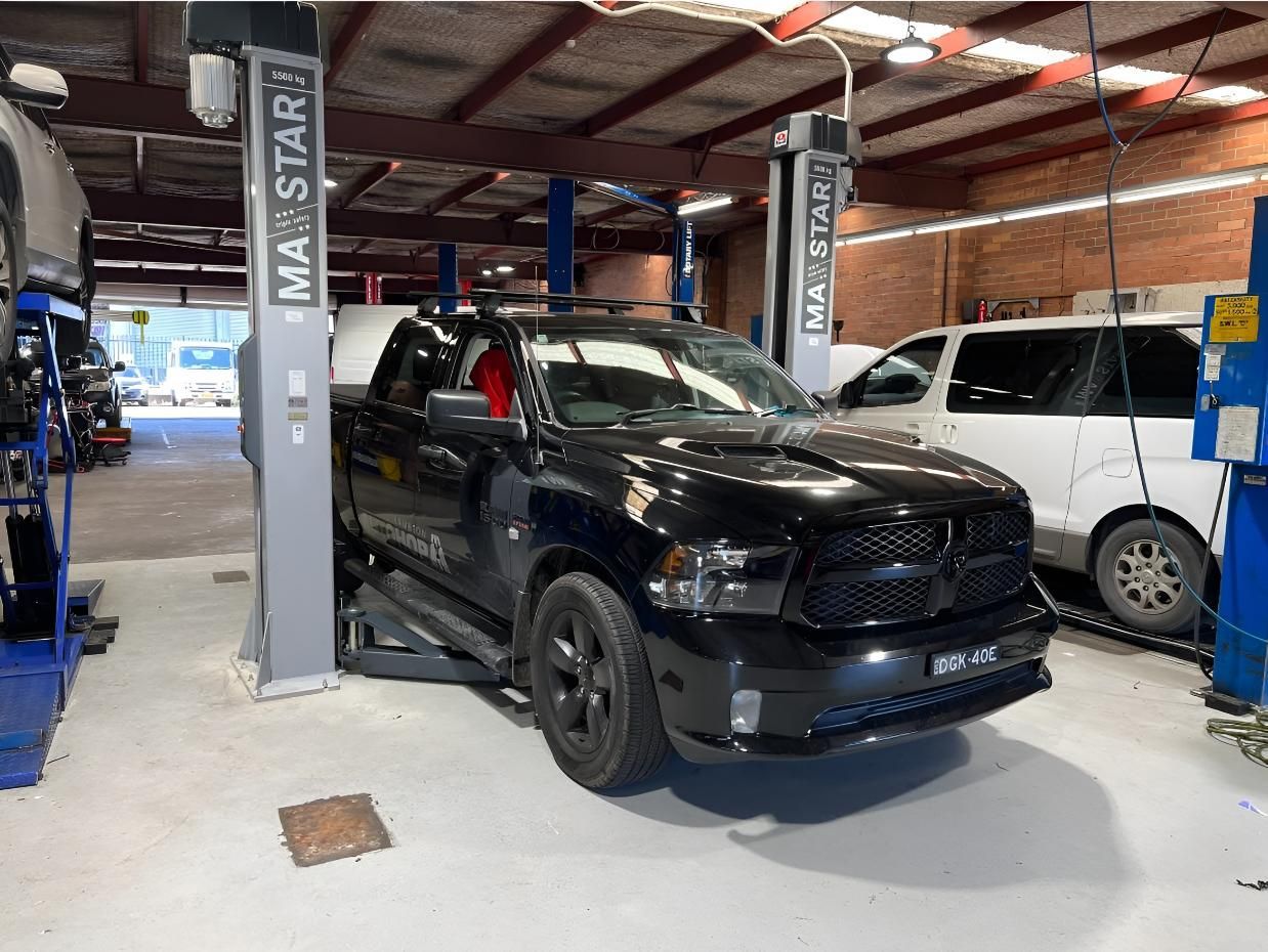 Black Pickup Truck on A Lift Inside a Garage, with A White Van and Tools Visible — AutoHop Lambton in Lambton, NSW
