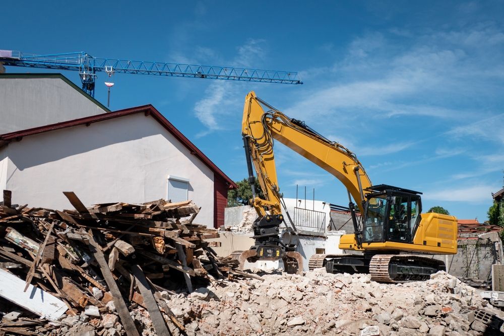 Yellow excavator demolishing a building; rubble, blue sky, and crane in the background.