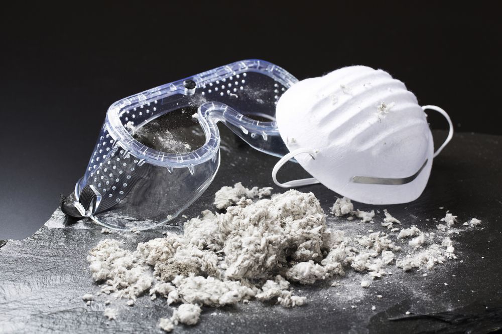 Protective gear with asbestos: goggles, mask, and pile of gray, fibrous material on a dark surface.