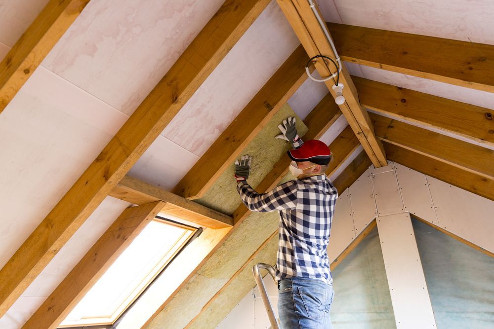 Person on a ladder installing insulation in an attic, wearing safety gear.