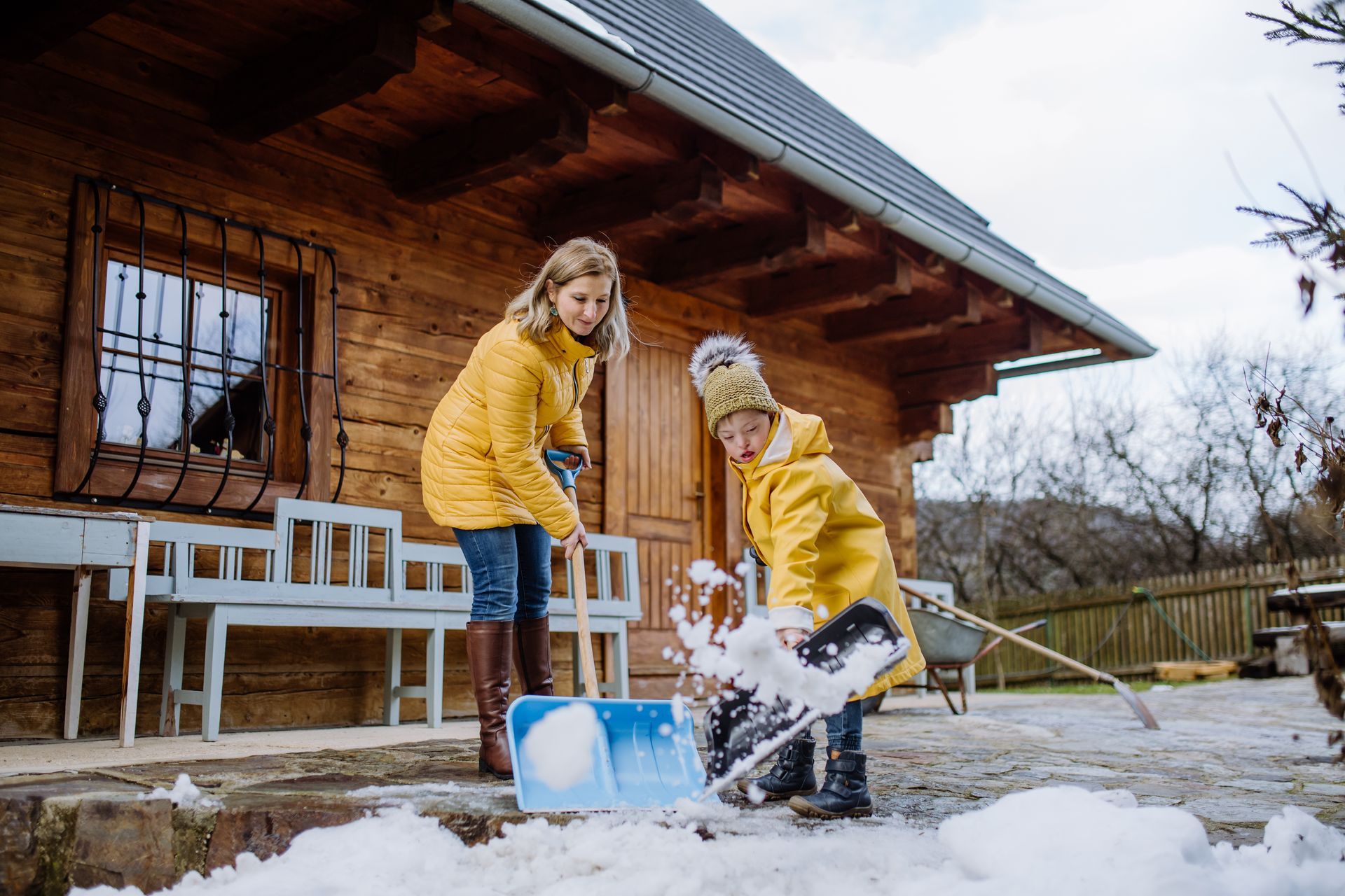 A woman and a child in yellow coats shovel snow off of a patio.