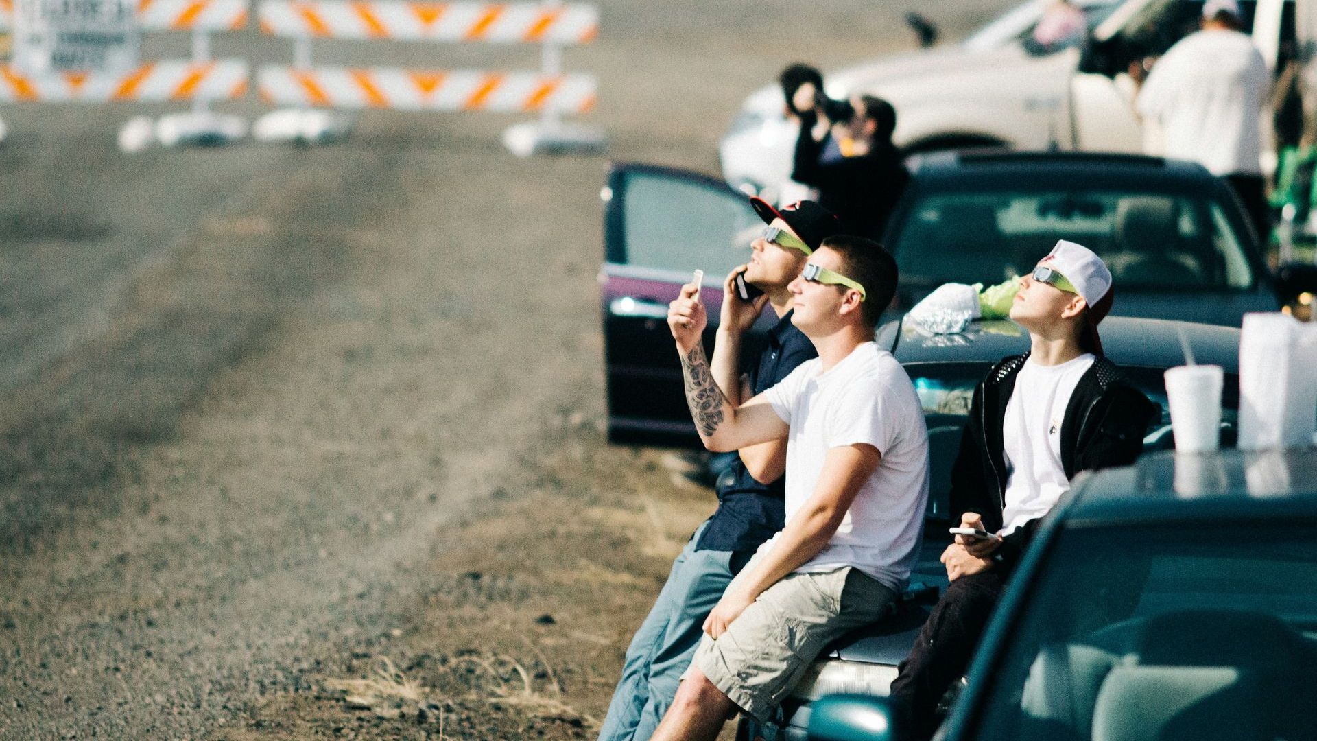 People sit on cars while wearing eclipse glasses. 