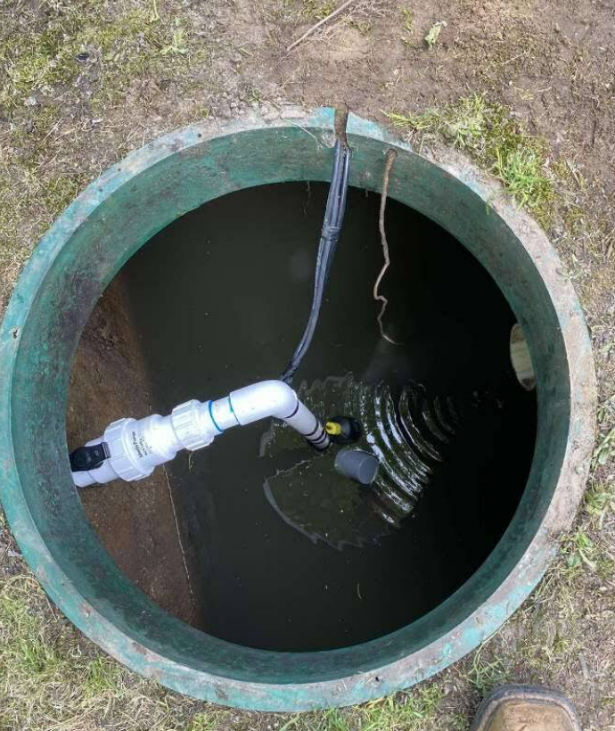 Open septic tank with water, pipes, and pump inside. Green concrete rim.