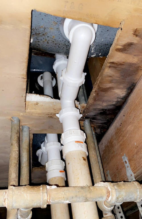 Plumbing pipes under a sink, white and beige, inside a wall cavity.