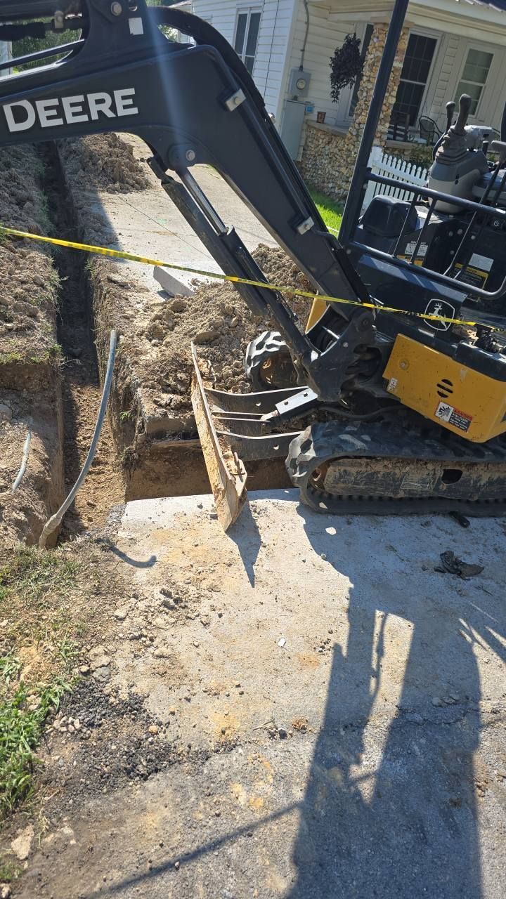A John Deere excavator digging a trench alongside a sidewalk in front of a house.