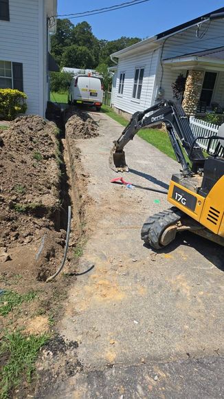 Yellow excavator digging a trench next to a house, with a utility van in the distance.