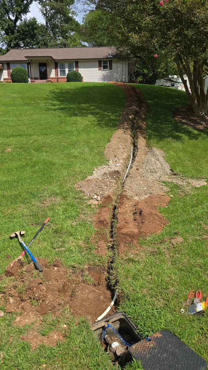 A trench dug in a grassy yard, with a house in the background. Tools lie nearby.