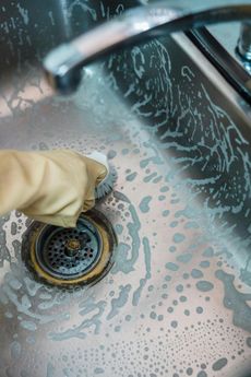 Person in gloves scrubbing a dirty sink drain with a brush.
