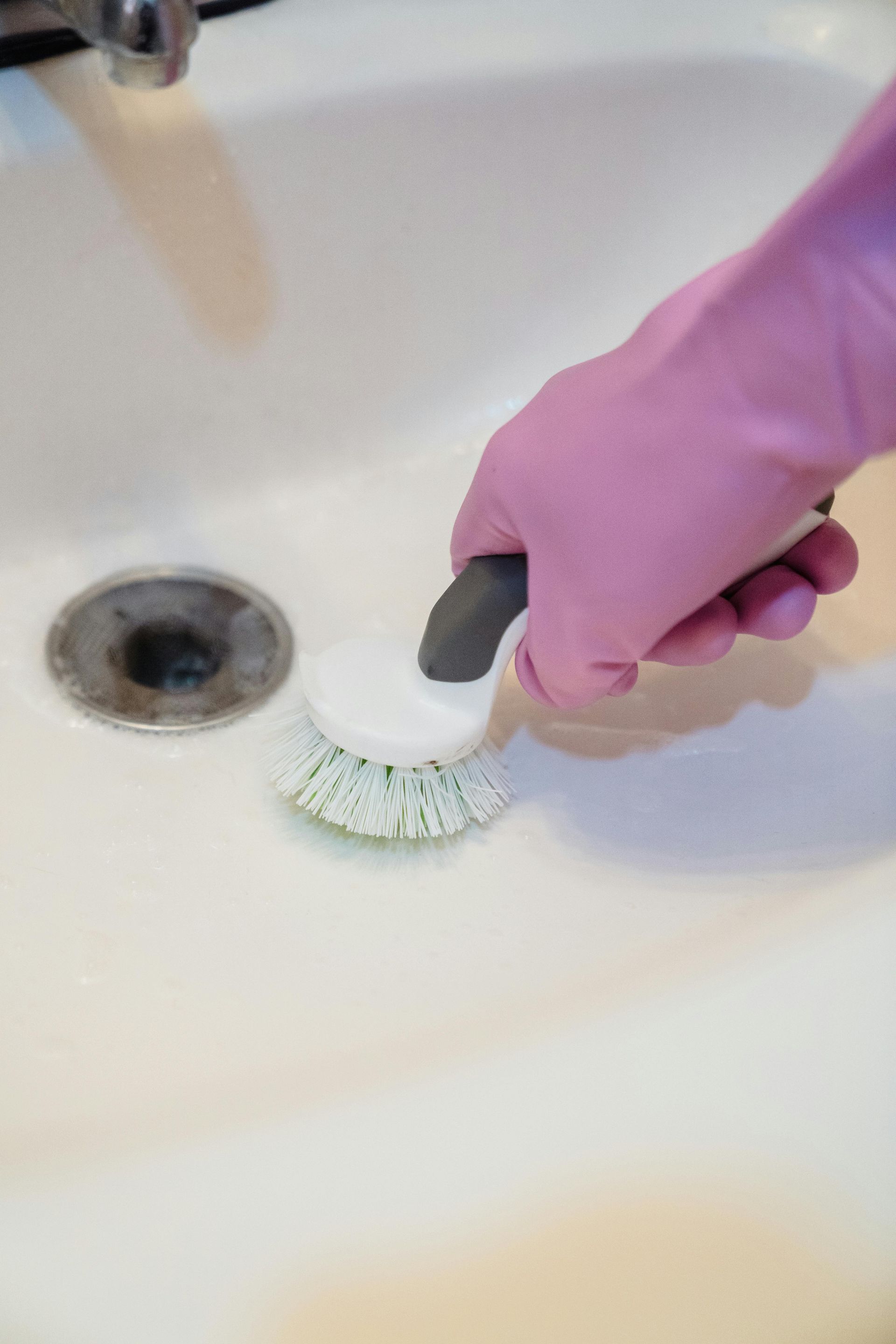 Pink-gloved hand scrubs a white sink with a round brush near a drain.