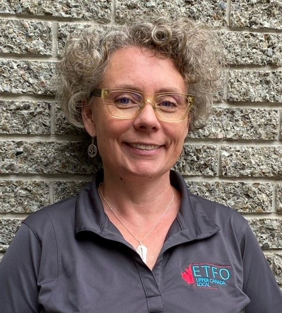 A woman wearing glasses and a gray shirt is smiling in front of a brick wall.