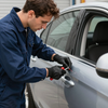 A worker in blue coveralls and black gloves uses a red tool to touch up a paint scratch on a silver car door.