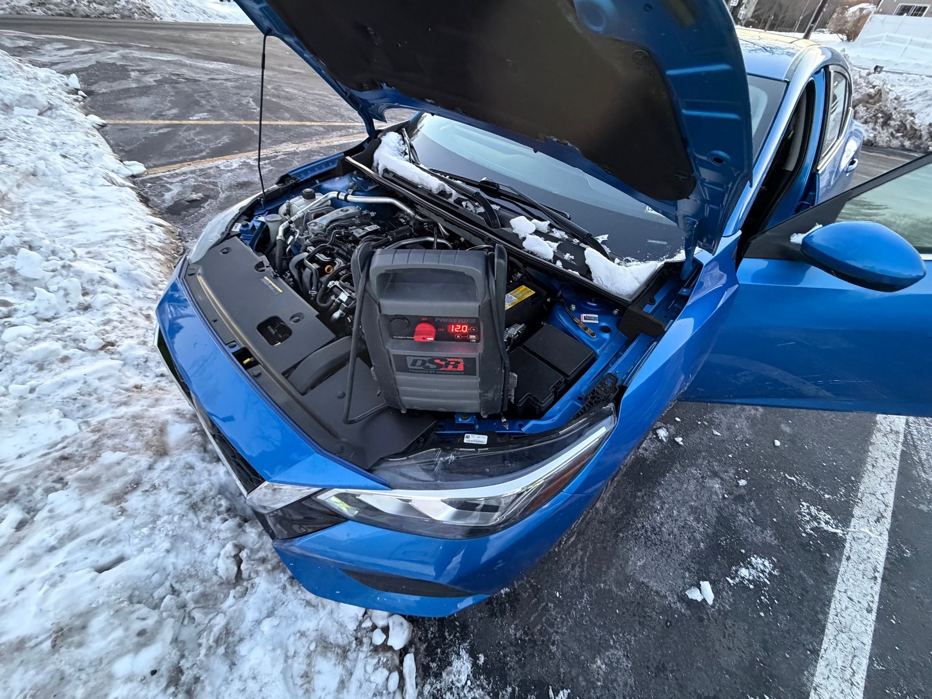 Blue car with hood open, showing the engine bay and battery in a snowy parking lot.