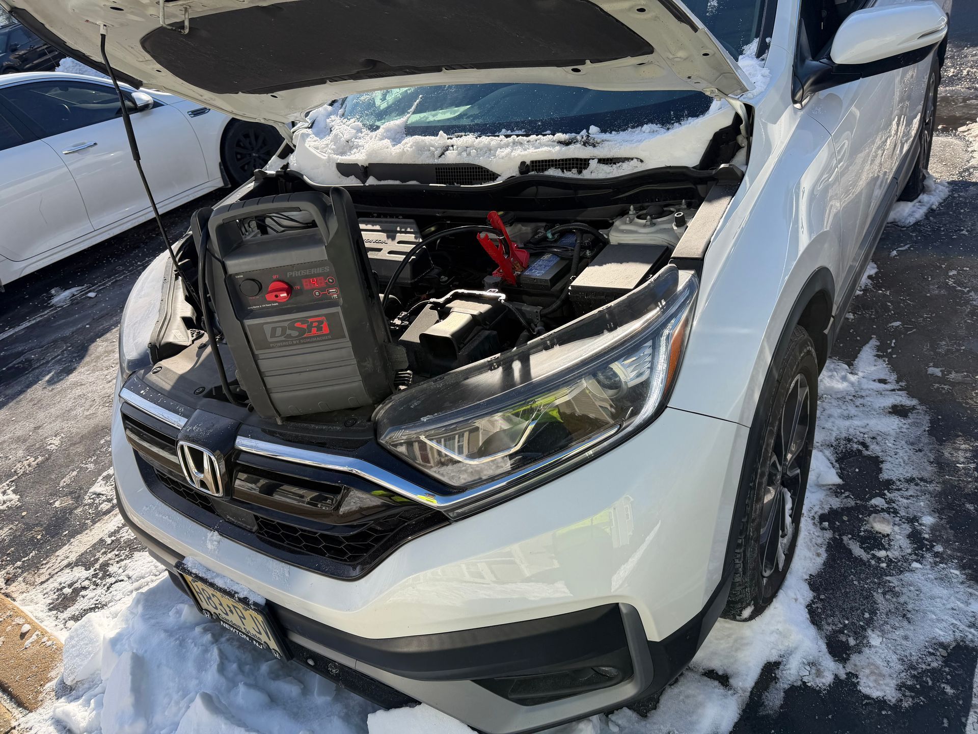 Silver Honda with hood open and jumper pack connected in snowy parking lot