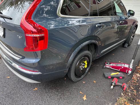 Gray SUV with rear wheel removed, sitting on a jack next to a red floor jack on a wet roadside