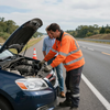 A road assistance worker in an orange high-visibility jacket examines the engine of a car on the side of a highway.