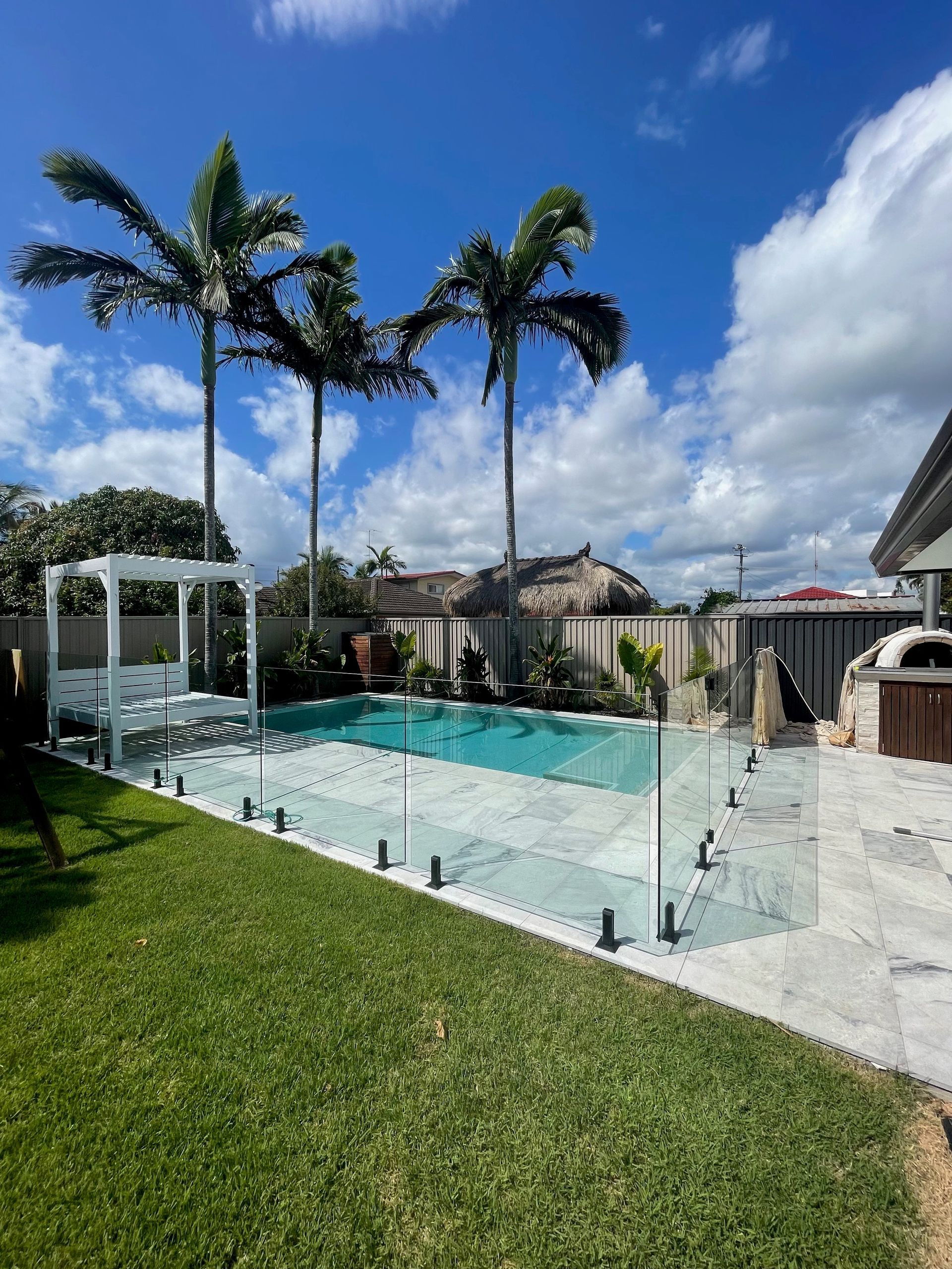 Backyard pool with glass fence, palm trees, and lawn on a sunny day.