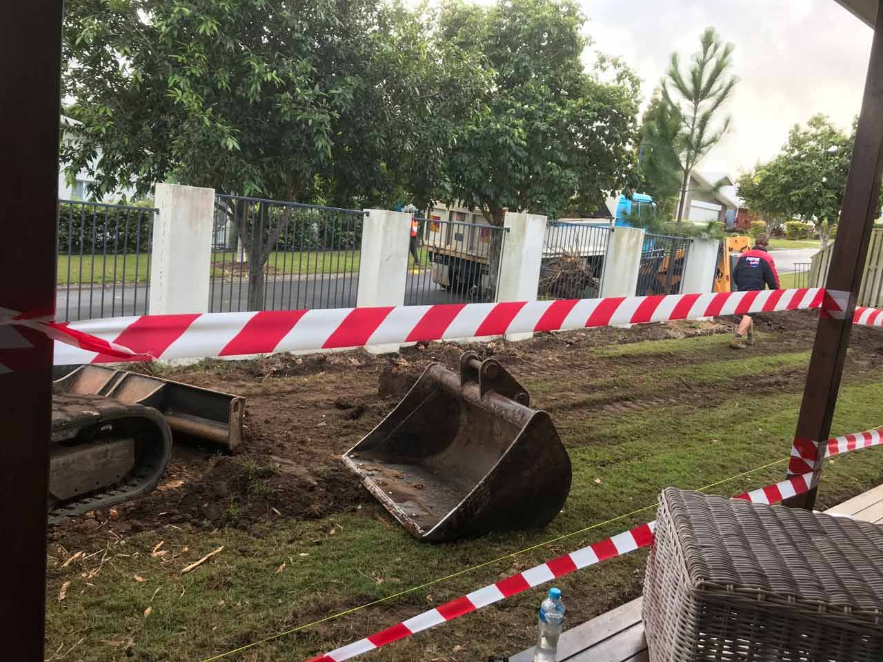Excavator bucket on muddy ground behind red and white caution tape. In a yard next to a fence.