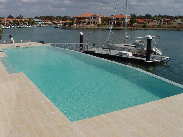 A rectangular swimming pool with an infinity edge next to a dock with a boat, in a waterfront residential area.