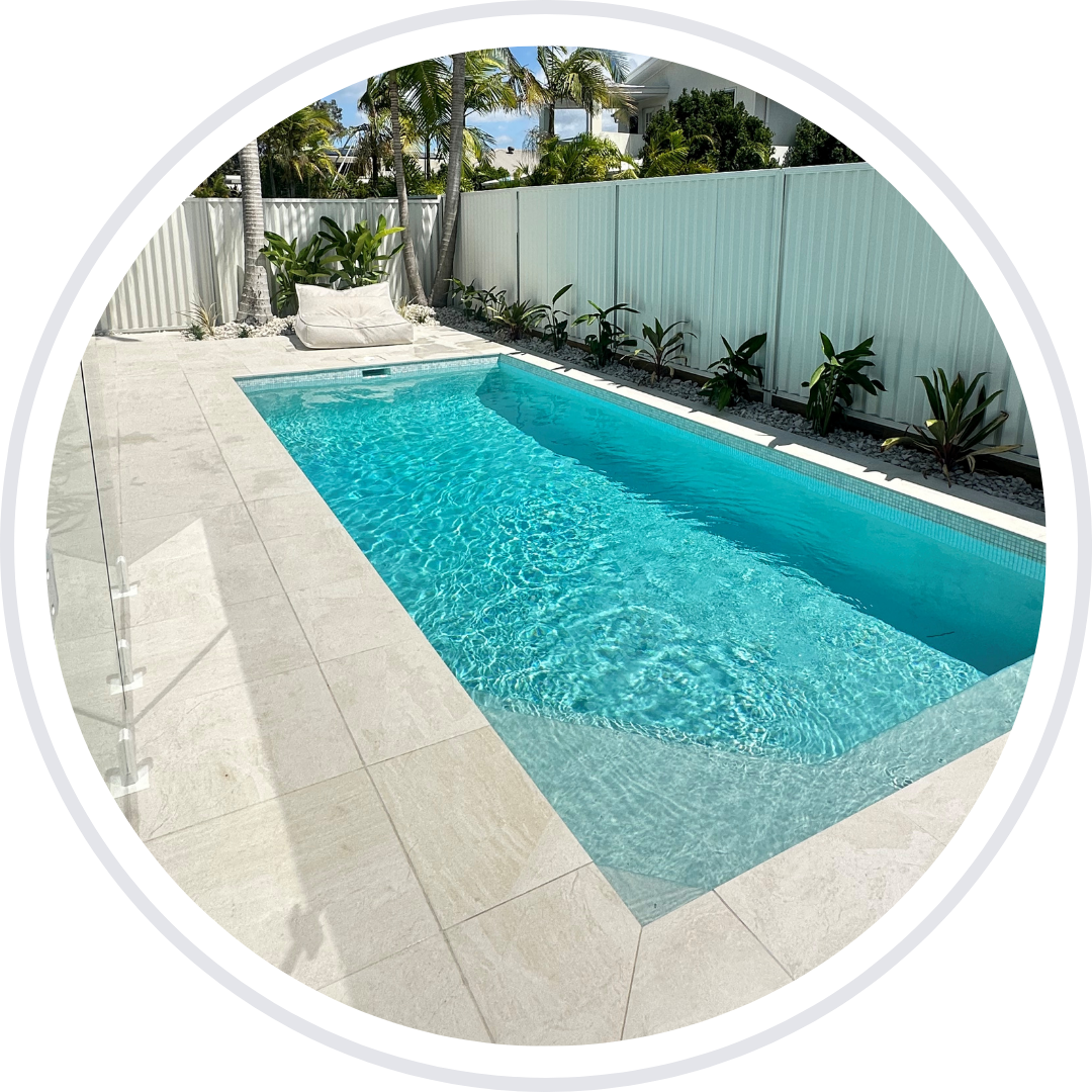Rectangular turquoise pool in a backyard with white tiling, fence, and tropical plants.