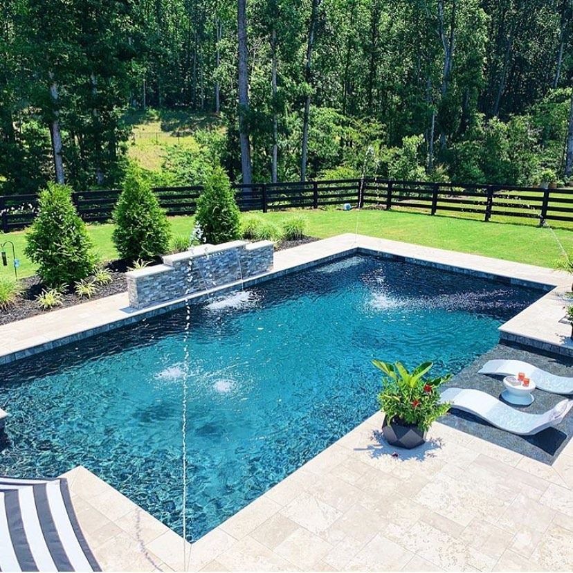 Rectangular pool with fountains, surrounded by stone and grass, backed by a forest and fence.