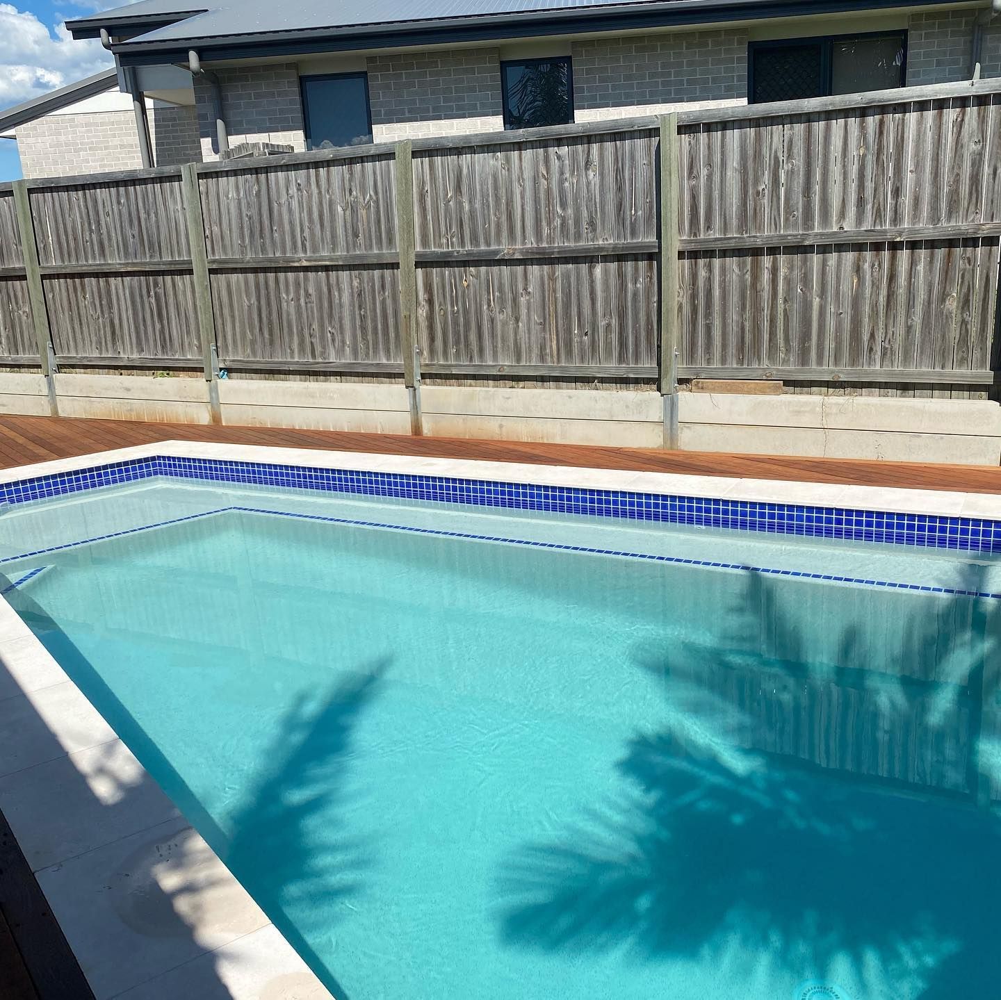 Swimming pool with blue water, surrounded by a wooden deck and a wooden fence. Palm tree shadow on water.