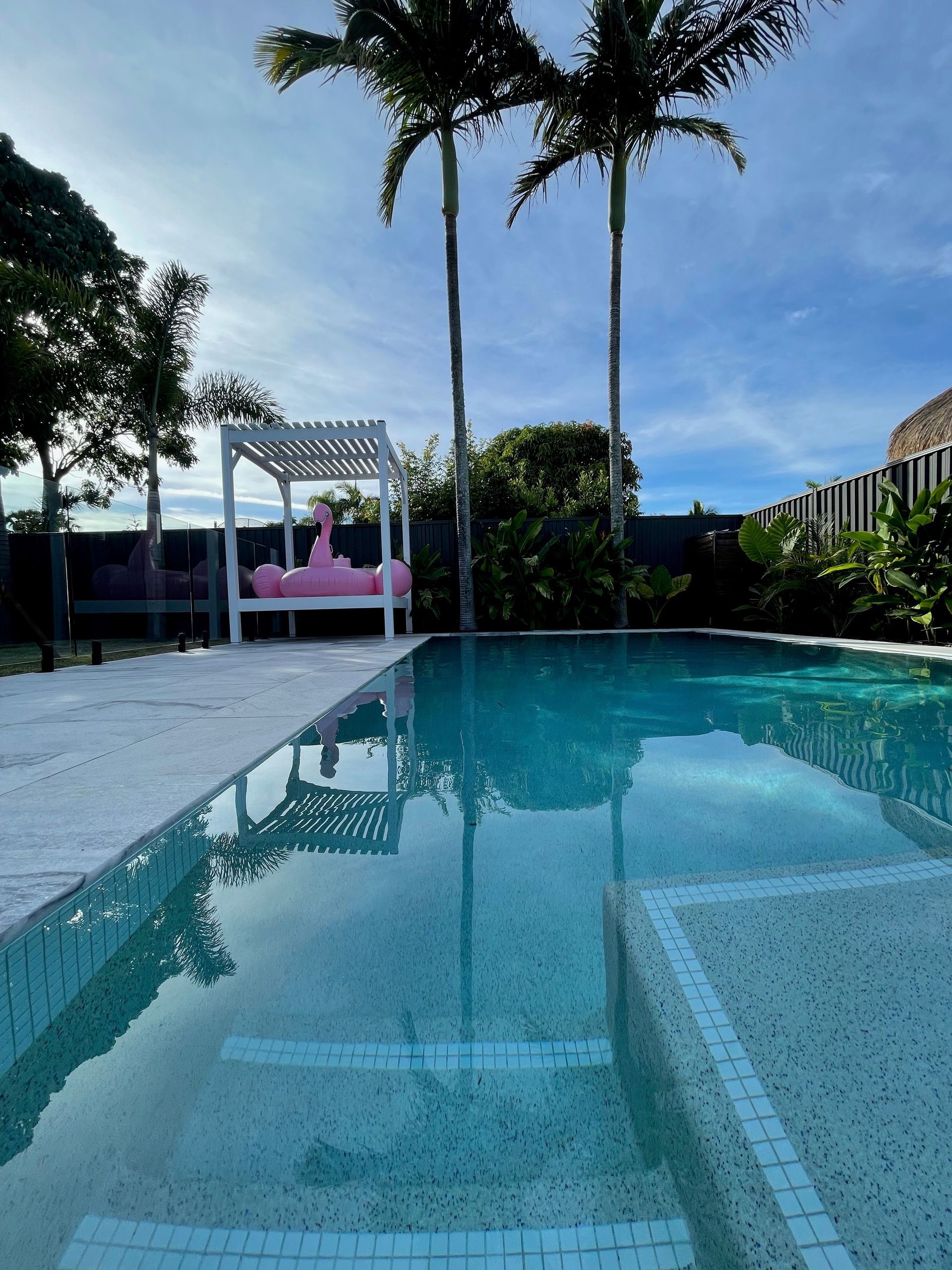 Swimming pool with clear water, white gazebo, and palm trees under a blue sky.