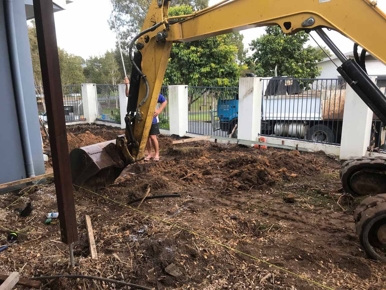 Yellow excavator digging in dirt near a fence, with a person standing nearby.
