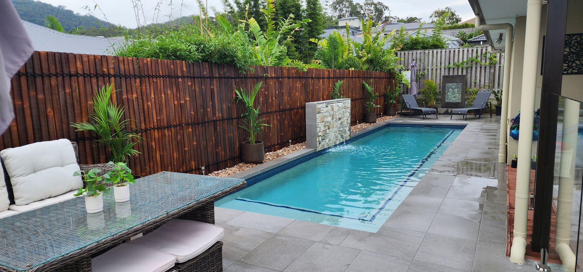 A rectangular swimming pool with a decorative stone wall in a yard. The pool is surrounded by a bamboo fence and lush greenery.