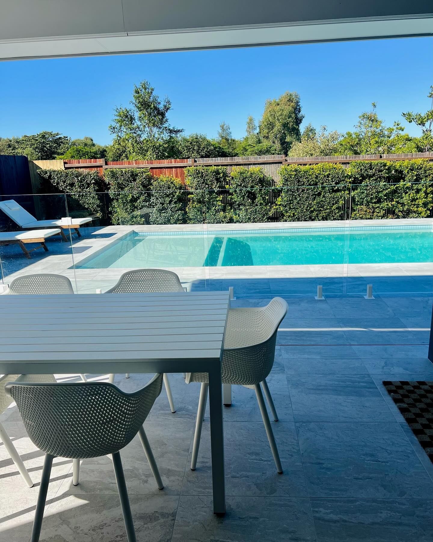 Outdoor patio with table and chairs overlooking a pool and greenery under a bright blue sky.