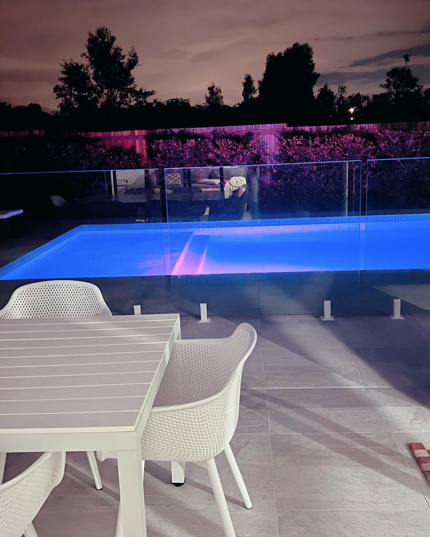 Outdoor pool with blue lighting, white furniture, and a fountain at twilight.