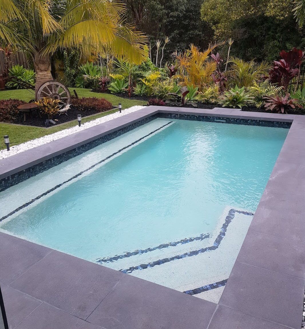 Rectangular pool with steps. Landscaped yard with palm tree, wheel, and greenery. Grey tile surround.