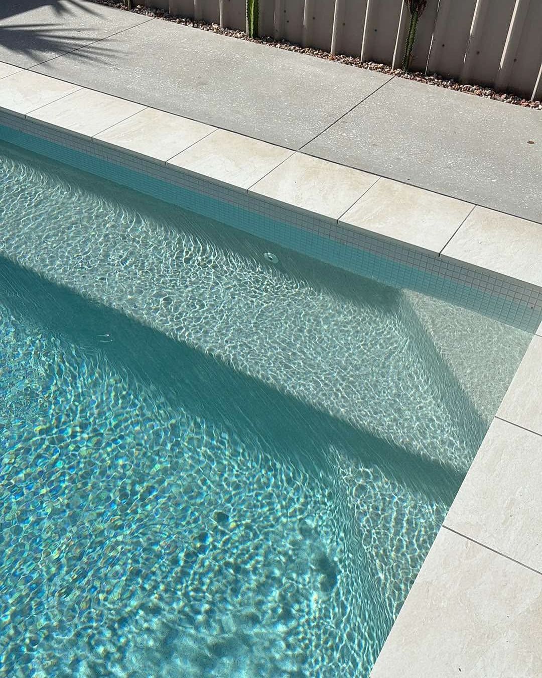 A rectangular swimming pool with steps, surrounded by light-colored tiles, with clear, turquoise water.