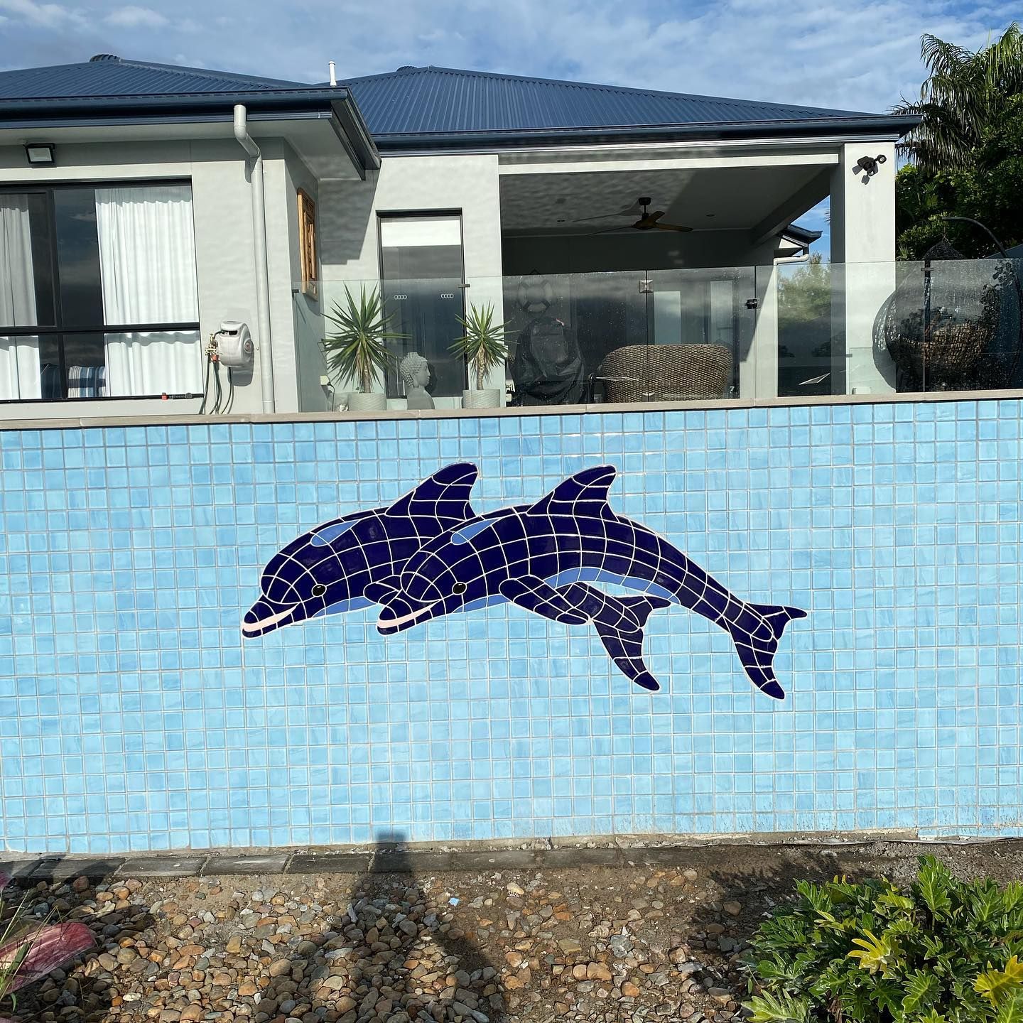 Two blue mosaic dolphins on a light blue tiled wall, beside a house with a blue roof.