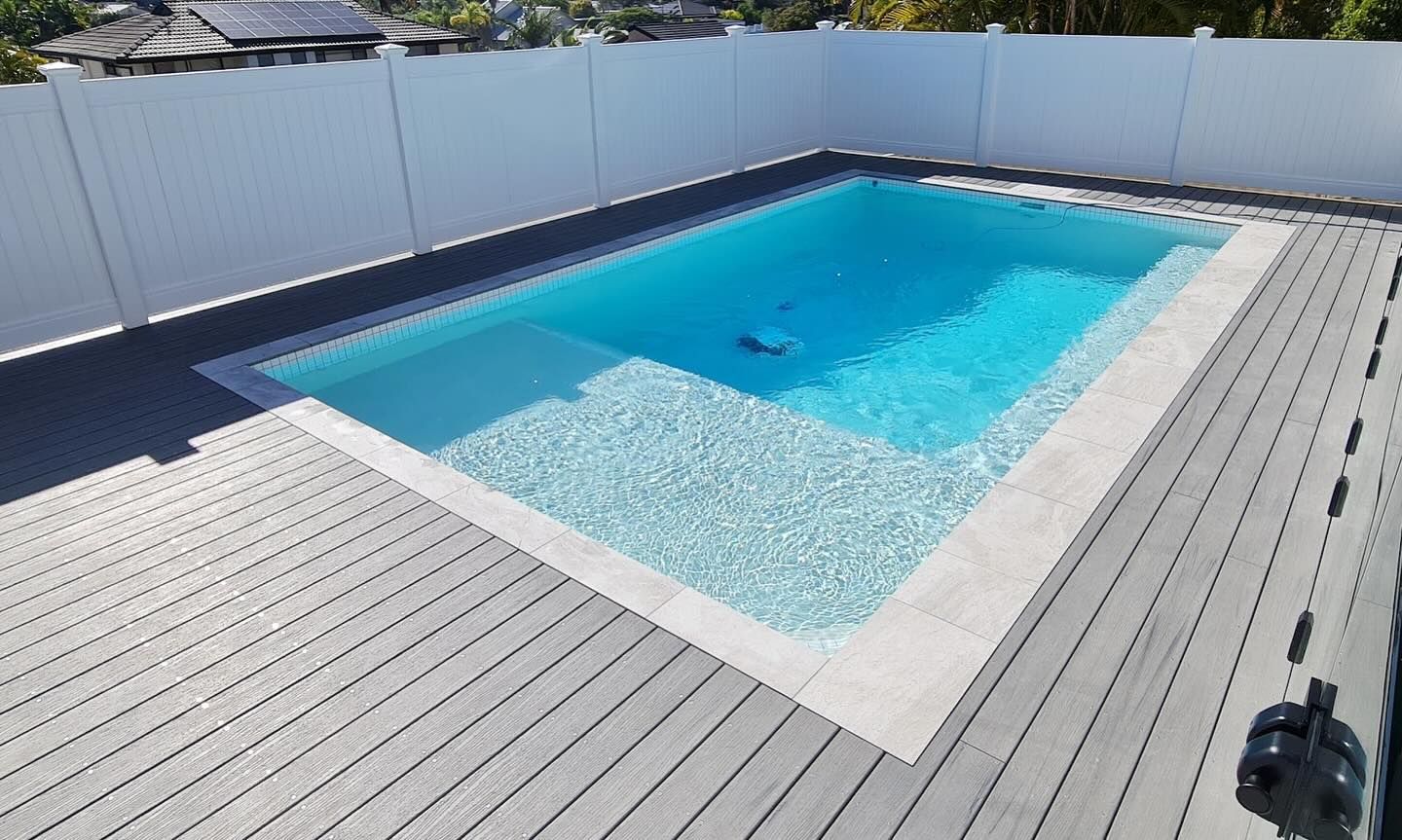 Rectangular pool with white interior, surrounded by gray decking and a white fence, on a sunny day.