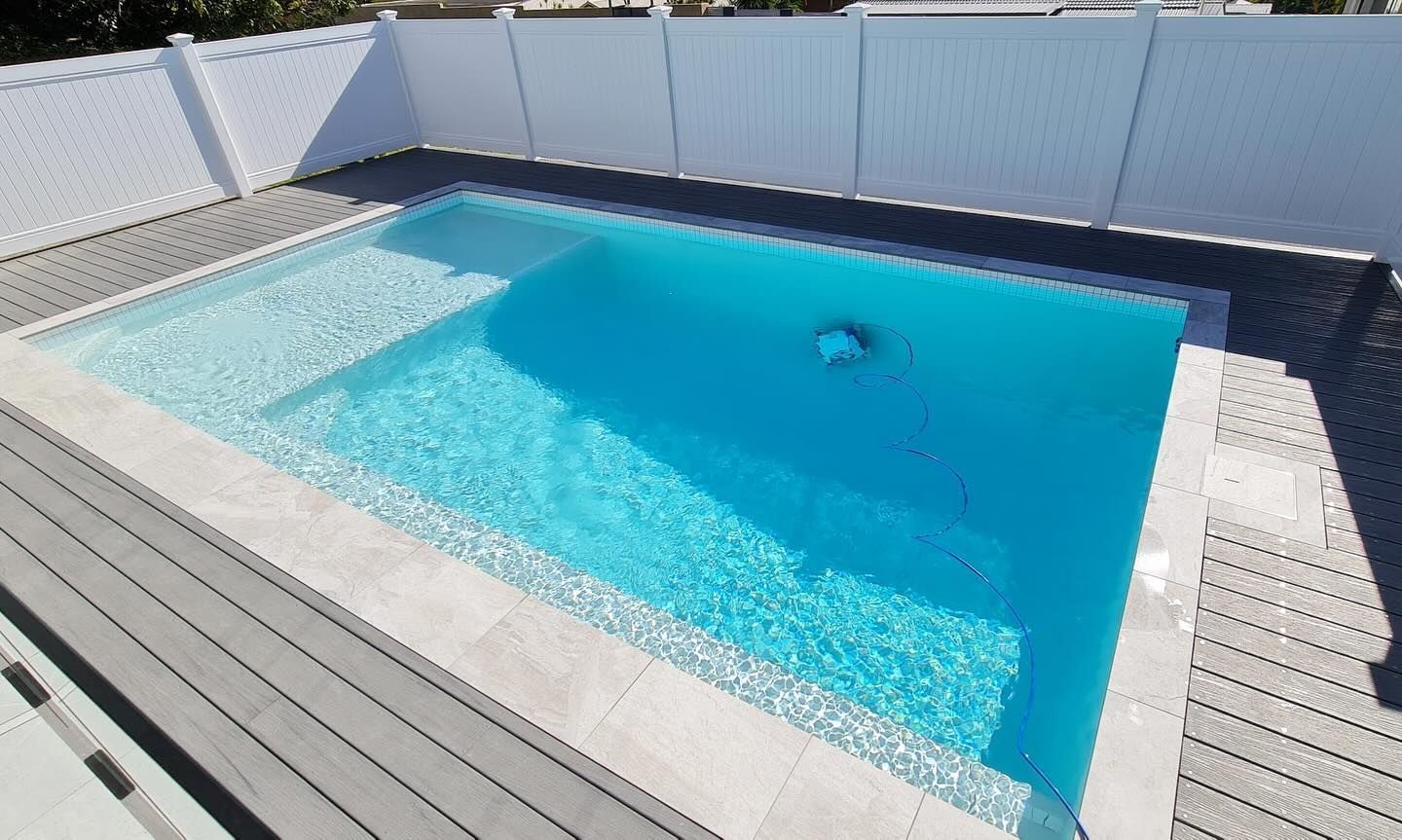 Rectangular pool with blue water, white steps, grey deck, and a white fence. Sunny outdoor setting.